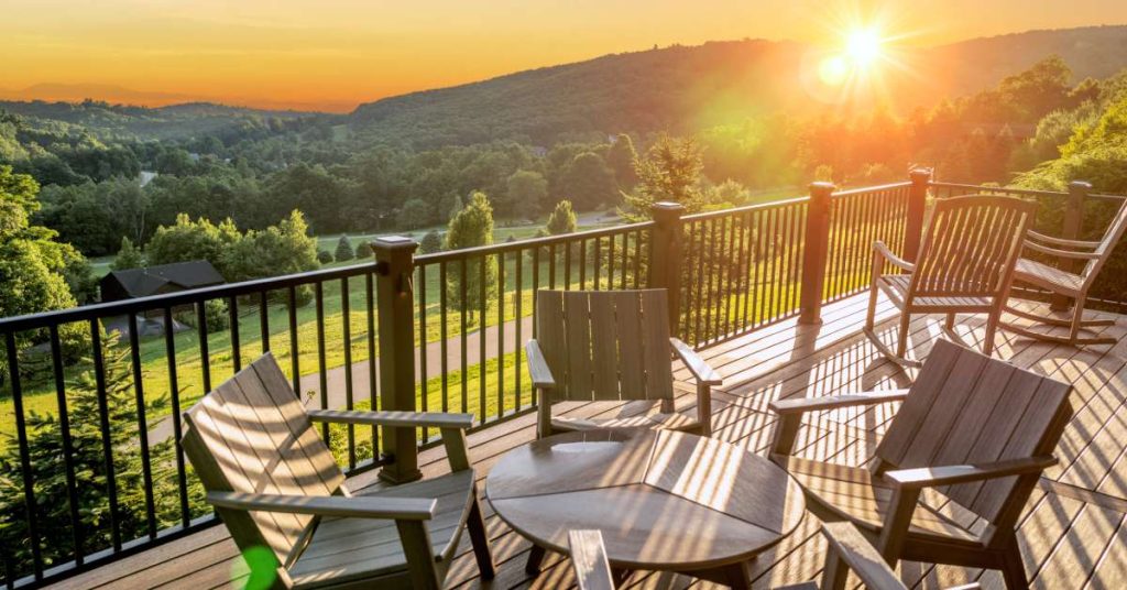 An outdoor deck overlooking a sunset. The balcony has black railings, brown plank floors, four chairs, and a table.